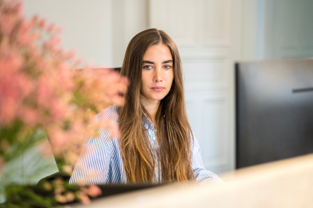 Eine EGIDO-Mitarbeiterin mit langen, braunen Haaren sitzt an einem Schreibtisch und schaut direkt in die Kamera. Im Vordergrund sind verschwommene pinke Blumen zu sehen, während der Hintergrund eine helle, minimalistische Büroatmosphäre zeigt.