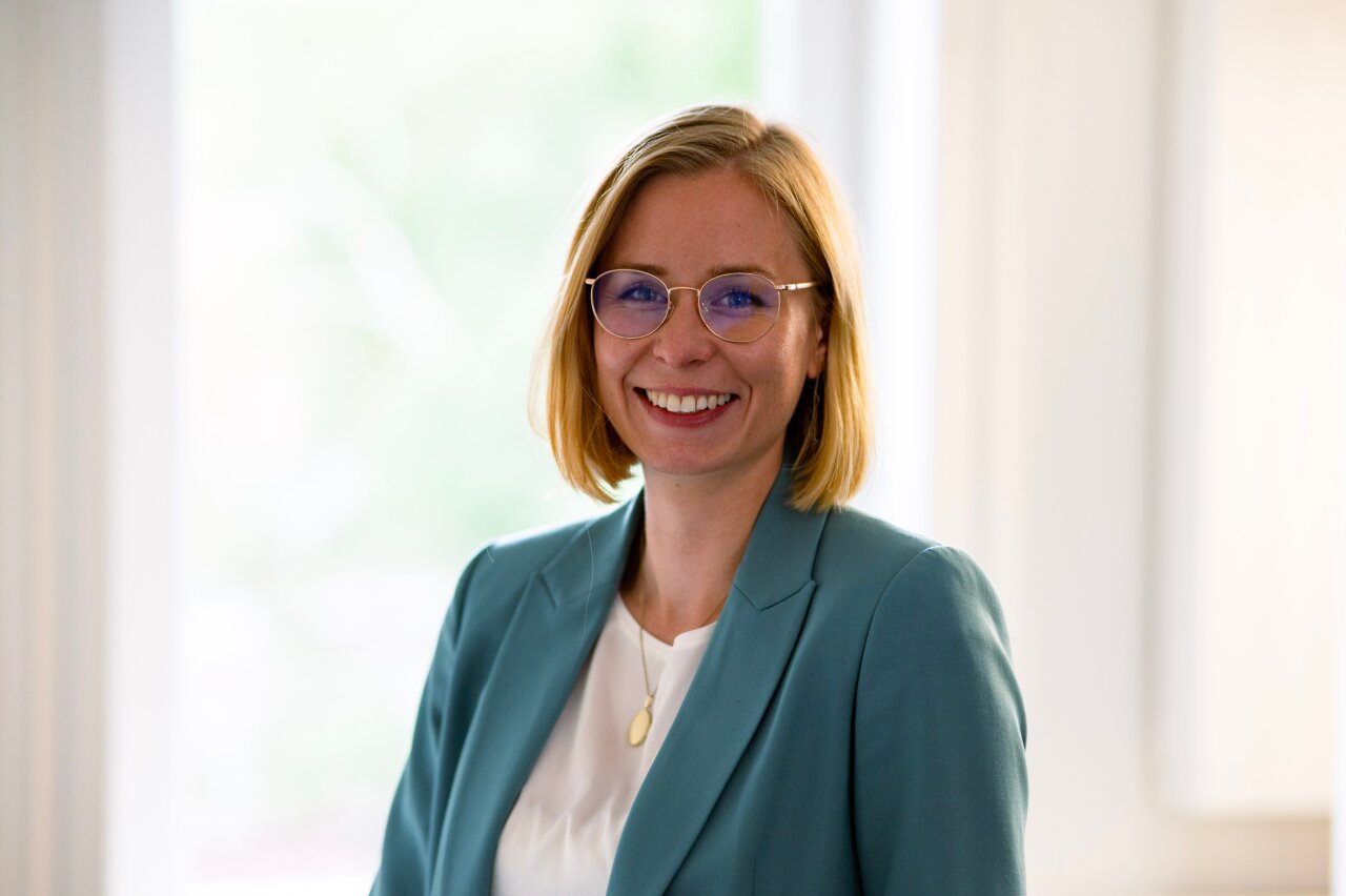 Eine lächelnde EGIDO-Mitarbeiterin mit blonden, kurzen Haaren trägt eine hellblaue Blazerjacke und eine weiße Bluse. Sie steht vor einem hellen Hintergrund mit einem weichen, unscharfen Licht.