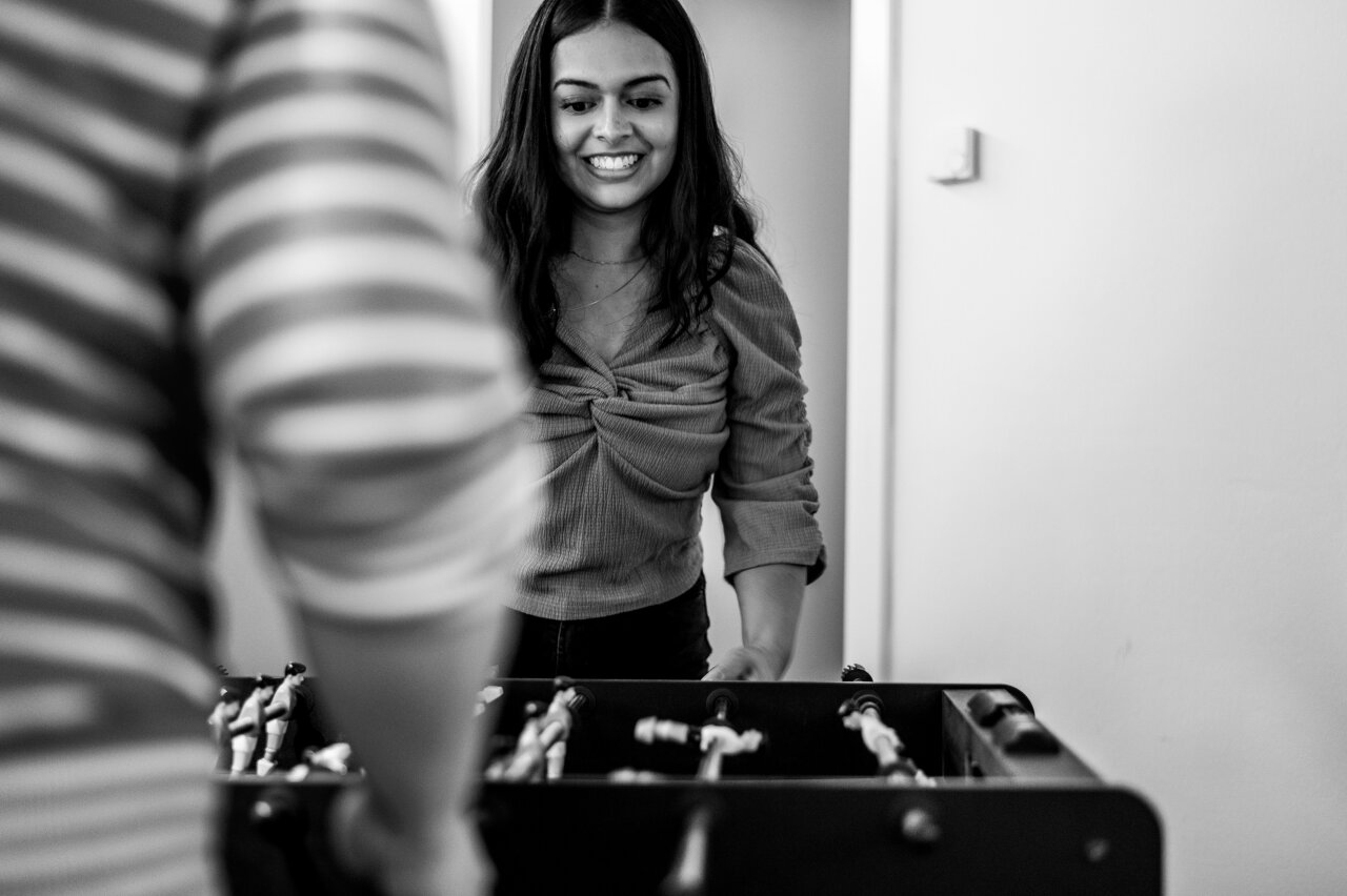 Eine EGIDO-Mitarbeiterin mit langen, dunklen Haaren lächelt, während sie an einem Tischfußballspiel steht. Im Vordergrund sind die gestreiften Ärmeln einer anderen Person sichtbar, während sie ebenfalls am Tischfußballspiel teilnehmen.