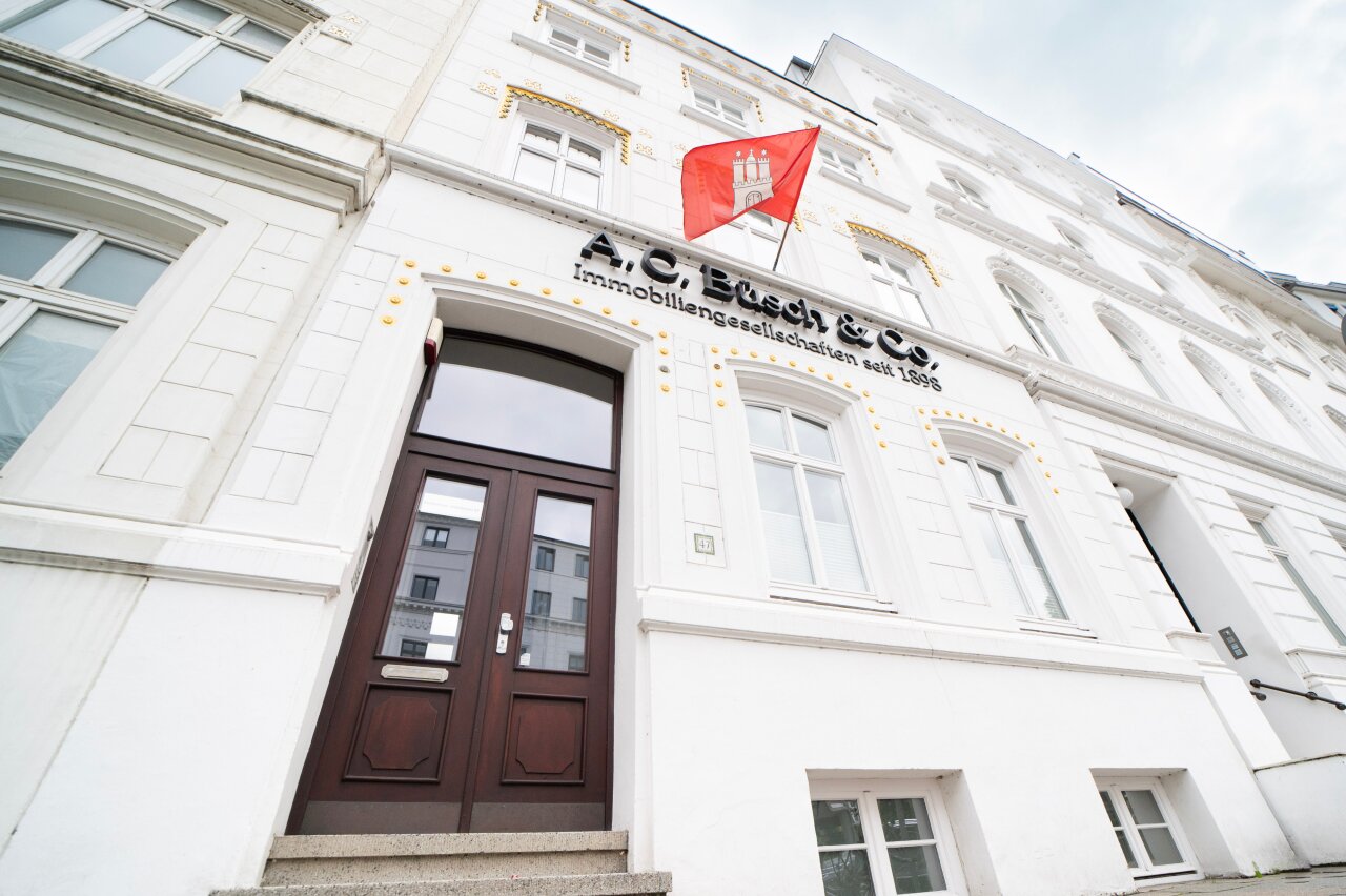 Das EGIDO-Gebäude hat eine helle, weiße Fassade mit goldenen Akzenten und großen Fenstern. Über der Eingangstür hängt eine rote Fahne mit dem Wappen Hamburgs. An der Fassade ist der Schriftzug 