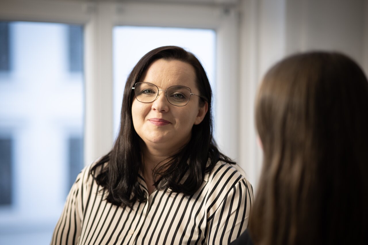 Eine EGIDO-Mitarbeiterin mit langen, glatten, schulterlangen Haaren und einer gestreiften Bluse sitzt in einem hellen Raum. Sie blickt lächelnd in die Kamera, während eine andere Person im Hintergrund leicht unscharf zu sehen ist.