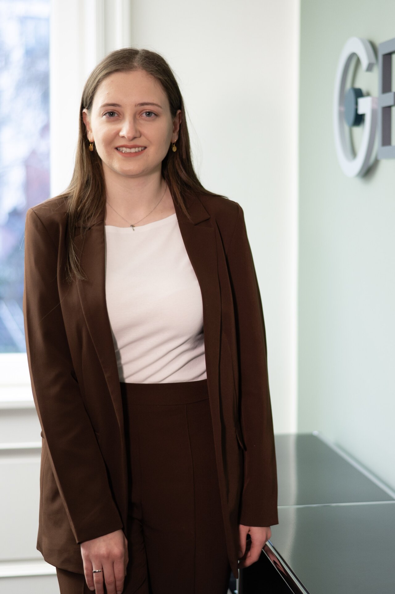 Eine EGIDO-Mitarbeiterin mit langen, braunen Haaren trägt einen braunen Anzug und ein weißes Oberteil. Sie steht in einem modernen Raum mit einem Fenster und einer grauen Oberfläche im Hintergrund.