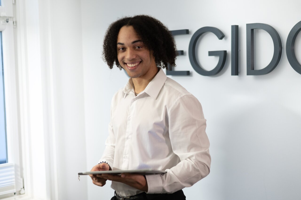 Ein junger EGIDO-Mitarbeiter mit lockigem Haar lächelt und hält ein Tablet in der Hand. Im Hintergrund sind große, graue Buchstaben an der Wand zu sehen, die offenbar einen Namen oder eine Marke darstellen.