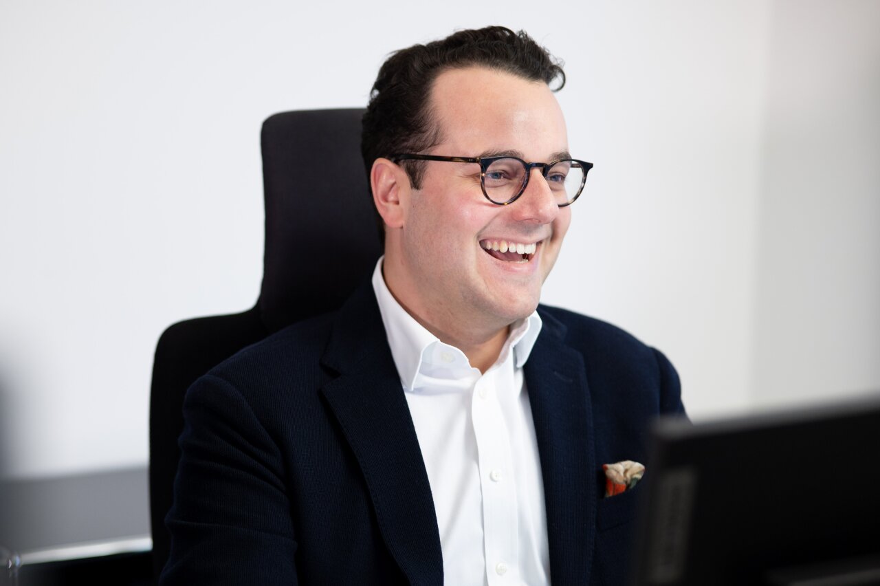 Ein lächelnder EGIDO-Mitarbeiter trägt eine Brille und einen dunkelblauen Blazer. Er sitzt an einem Schreibtisch und schaut auf einen Computerbildschirm, während er fröhlich aussieht.