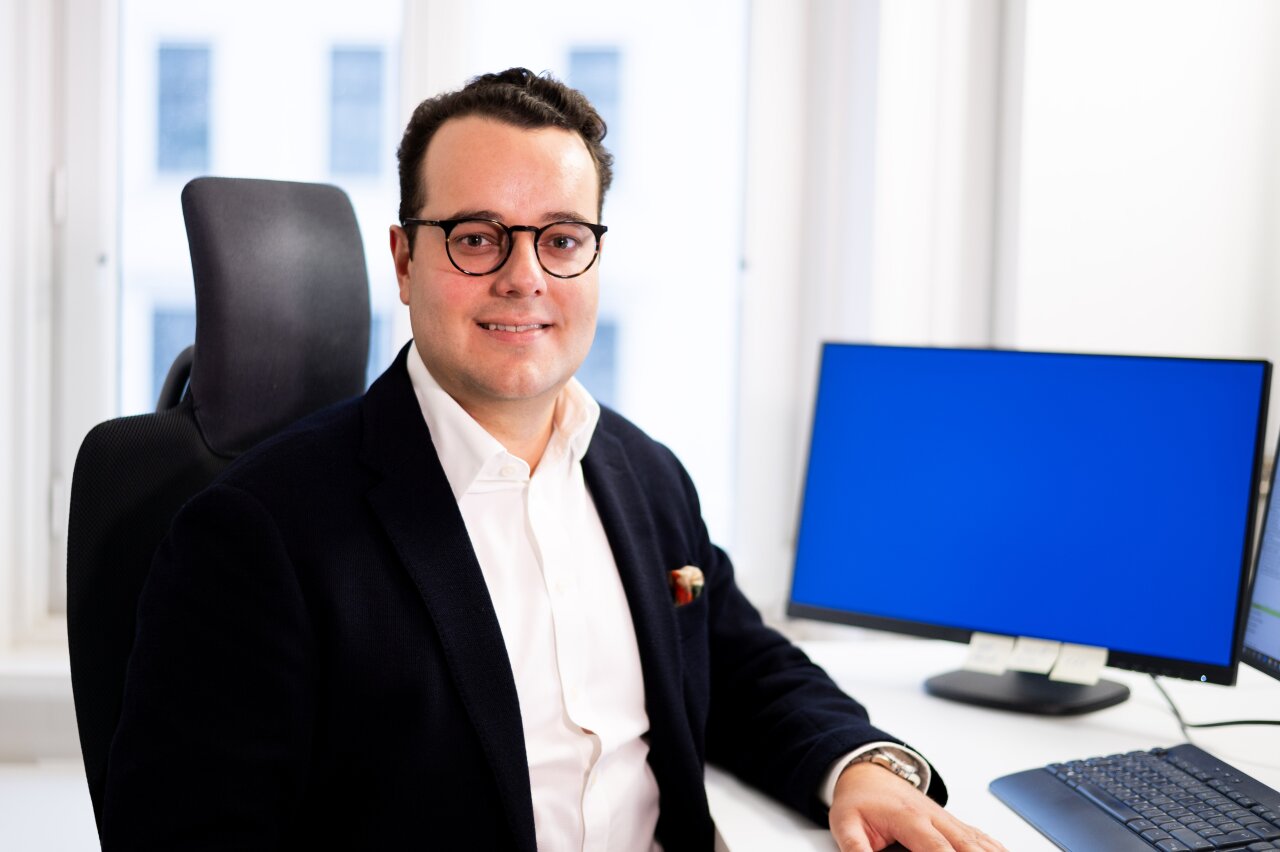 Ein EGIDO-Mitarbeiter mit Brille sitzt an einem Schreibtisch in einem modernen Büro. Er trägt einen dunklen Blazer über einem weißen Hemd und lächelt, während ein großer, blauer Computerbildschirm neben ihm sichtbar ist.