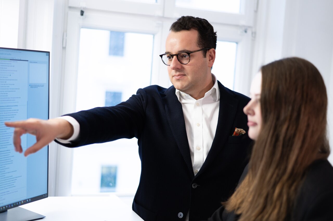 Ein EGIDO-Mitarbeiter mit Brille, der einen dunklen Blazer trägt, zeigt mit dem Finger auf einen Monitor, auf dem Text angezeigt wird. Neben ihm steht eine EGIDO-Mitarbeiterin mit langen, offenen Haaren, die aufmerksam zuhört.