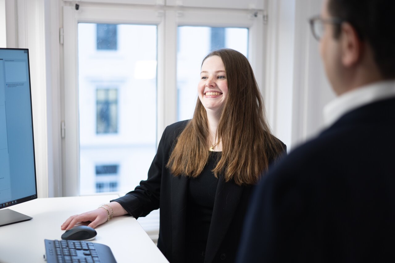 Eine junge EGIDO-Mitarbeiterin mit langen, braunen Haaren steht an einem Tisch und lächelt. Sie trägt einen schwarzen Blazer und schaut auf einen Bildschirm vor ihr, während eine Person im Hintergrund steht.