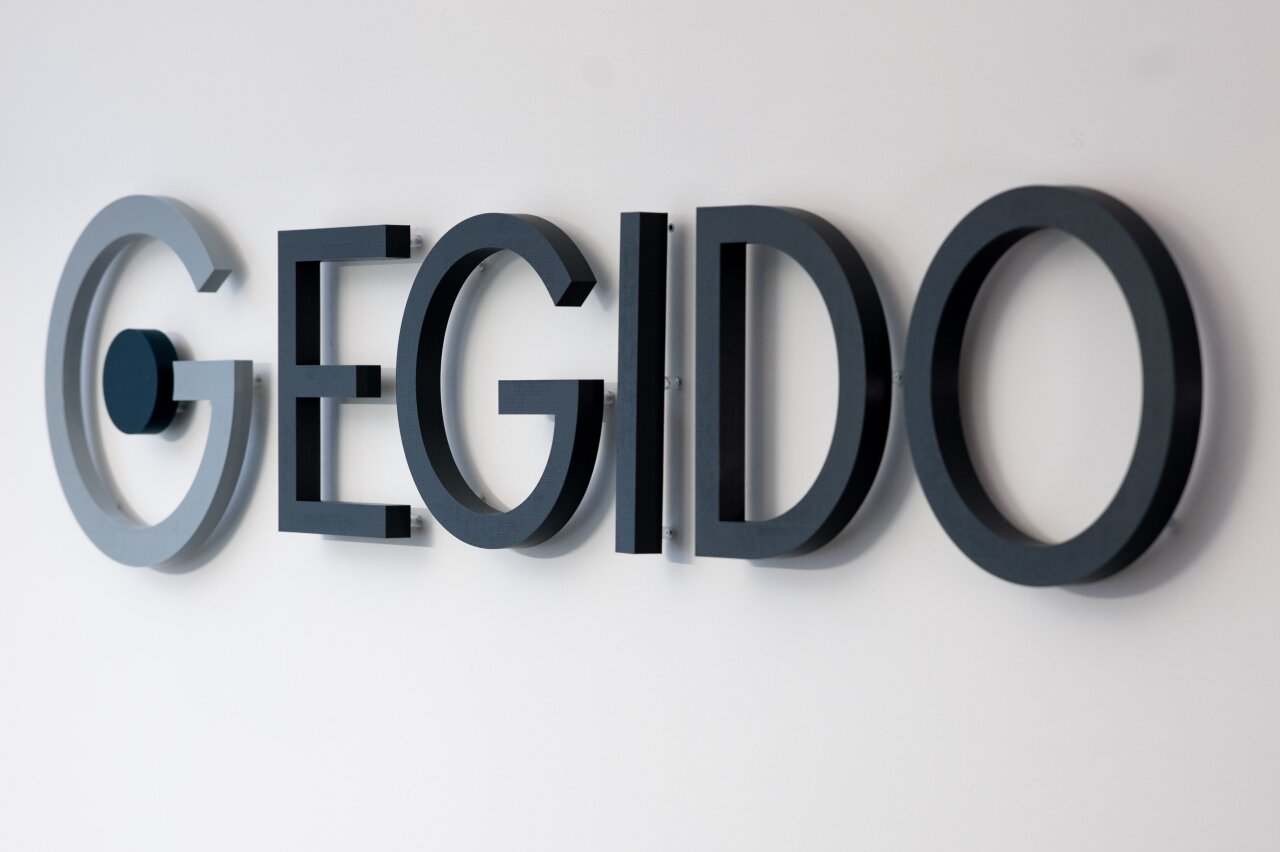 Der Schriftzug „EGIDO“ besteht aus großen, dreidimensionalen Buchstaben in verschiedenen Grautönen und ist an einer hellen Wand montiert.