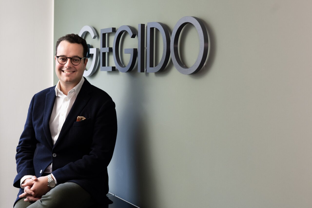 Ein lächelnder EGIDO-Mitarbeiter mit Brille sitzt auf einem Stuhl vor einer Wand mit dem Schriftzug 