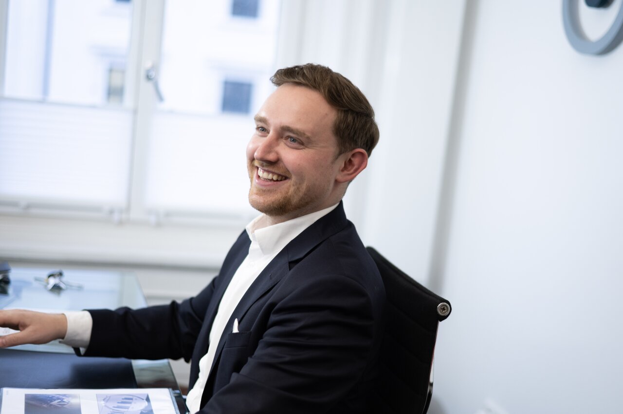 Ein lächelnder EGIDO-Mitarbeiter sitzt an einem Schreibtisch in einem modernen Büro. Er trägt einen dunkelblauen Anzug und hat die Haare nach hinten frisiert, während er auf ein Dokument schaut, das vor ihm liegt.