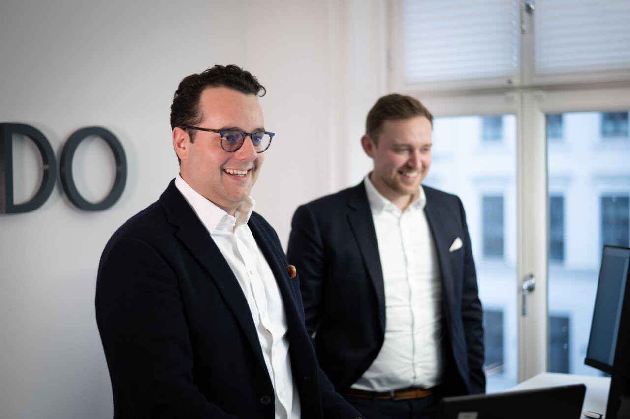 Zwei EGIDO-Mitarbeiter in Anzügen stehen an einem Tisch in einem Büro. Der EGIDO-Mitarbeiter links lächelt und trägt eine Brille, während der EGIDO-Mitarbeiter rechts ebenfalls lächelt und eine helle Hemdjacke trägt, beide scheinen sich über etwas am Computer zu freuen. Hinter ihnen ist ein großes Fenster und an der Wand steht der Schriftzug „EGIDO“.