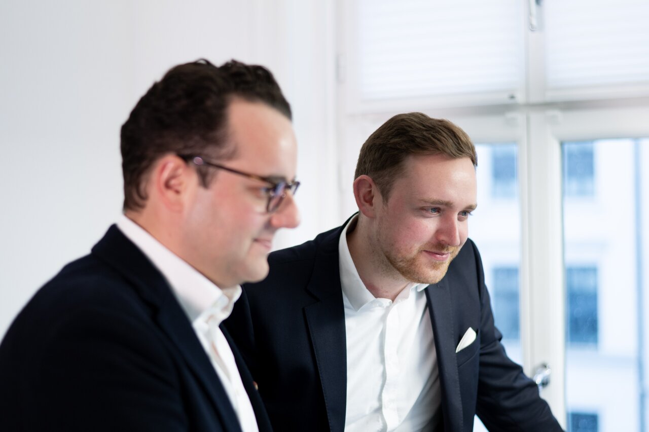 Zwei EGIDO-Mitarbeiter in Anzügen stehen in einem modernen Raum mit großen Fenstern. Einer von ihnen trägt eine Brille und schaut nachdenklich auf einen Bildschirm, während der andere lächelt und interessiert wirkt.