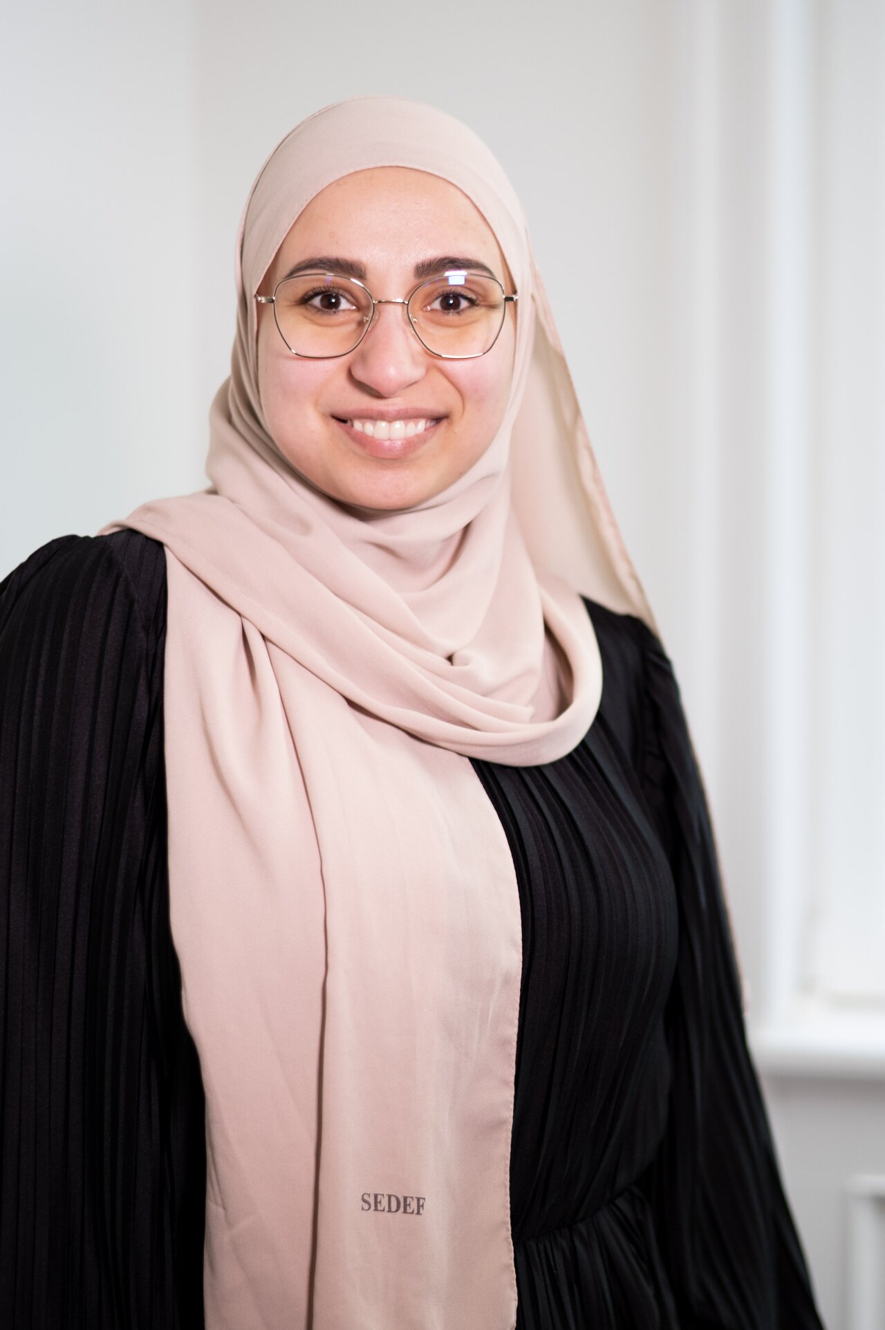 Eine junge EGIDO-Mitarbeiterin mit einem hellbeigen Hijab und einer schwarzen, plissierten Bluse lächelt freundlich in die Kamera. Sie trägt eine Brille und steht vor einem hellen Hintergrund, der das Bild gut beleuchtet.
