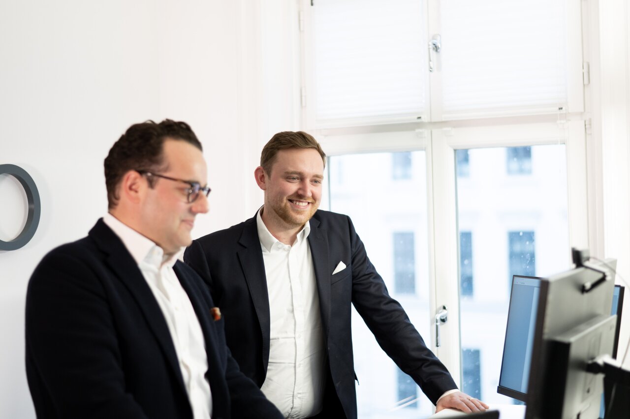 Zwei EGIDO-Mitarbeiter stehen in einem hellen Büroraum neben einem Computer und betrachten den Bildschirm. Der EGIDO-Mitarbeiter links trägt eine Brille und einen dunklen Blazer, während der EGIDO-Mitarbeiter rechts in einem schwarzen Anzug lächelt. Durch das Fenster ist eine urbane Umgebung zu sehen.