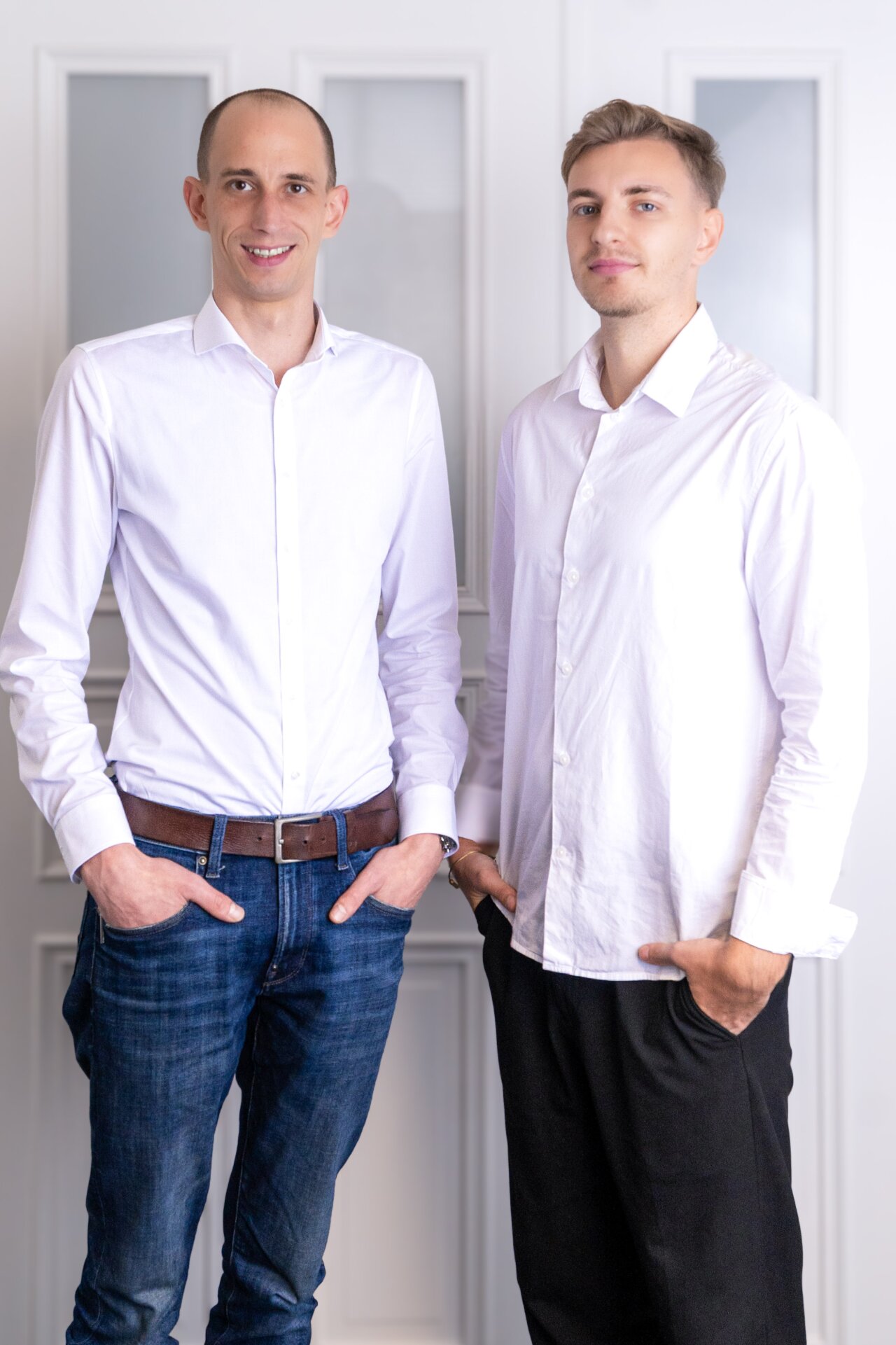 Zwei EGIDO-Mitarbeiter stehen nebeneinander vor einer hellen Wand. Beide tragen weiße Hemden, der auf der linken Seite kombiniert mit blauen Jeans und einem braunen Gürtel, der andere mit schwarzen Hosen. Sie lächeln und haben jeweils eine Hand in der Hosentasche.