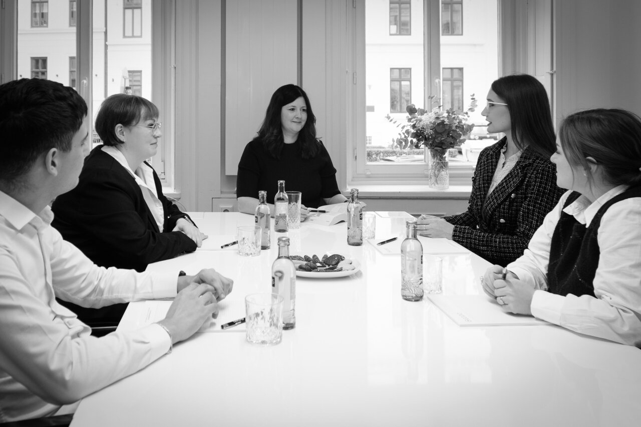 Fünf EGIDO-Mitarbeiter sitzen an einem Tisch in einem hellen Besprechungsraum. Auf dem Tisch stehen Wasserflaschen, Getränke in Gläsern und ein Teller mit Snacks. Im Hintergrund sind Fenster mit Blick auf eine grüne Pflanzenarrangement sowie Gebäude zu sehen.
