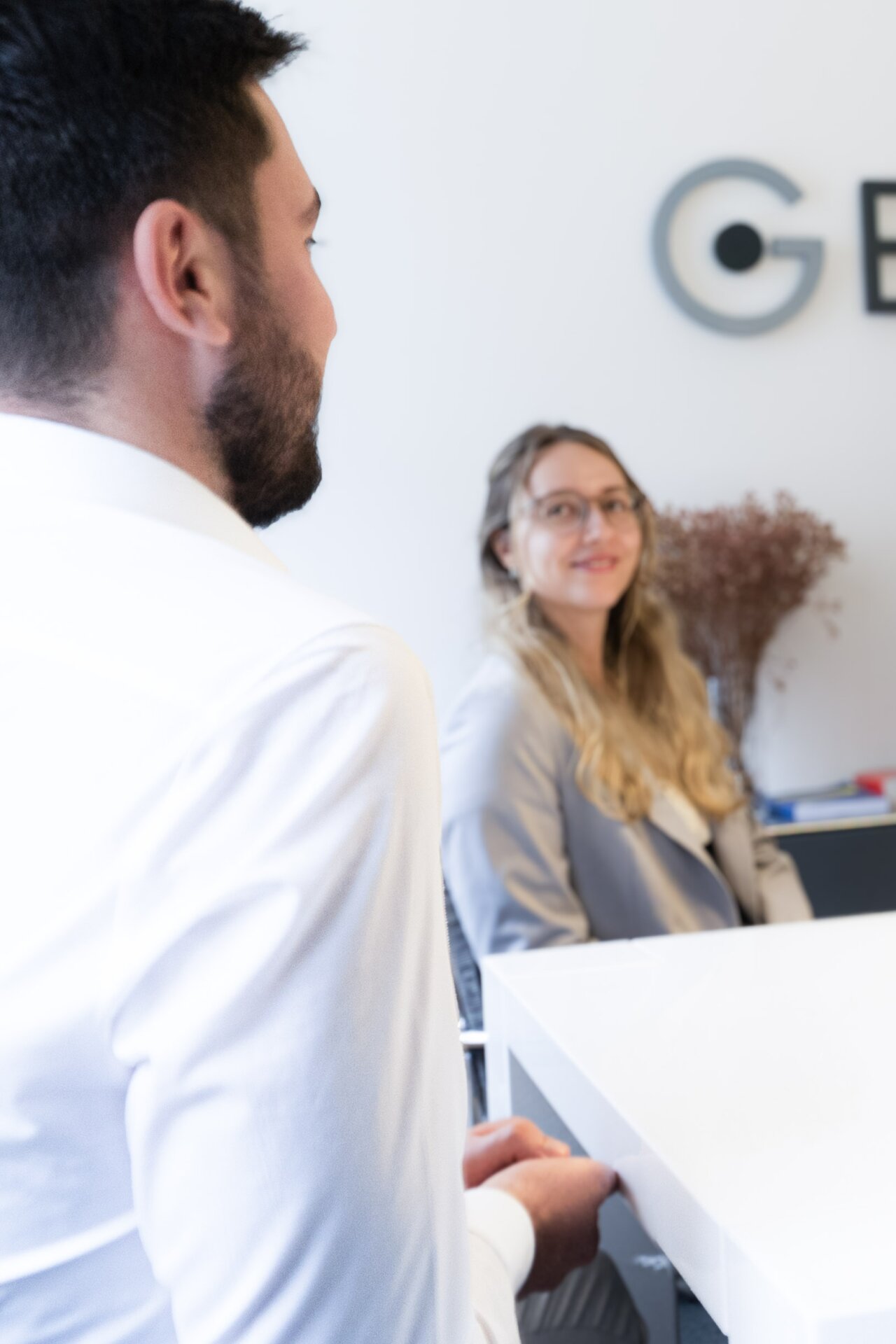 Ein EGIDO-Mitarbeiter in einem weißen Hemd sitzt mit dem Rücken zur Kamera und schaut auf eine EGIDO-Mitarbeiterin, die ihm gegenüber sitzt. Die √in trägt eine graue Jacke und hat lange, lockige Haare; sie lächelt leicht und scheint an dem Gespräch interessiert zu sein. Im Hintergrund ist ein helles Büro mit einer Pflanze und Büromaterialien.