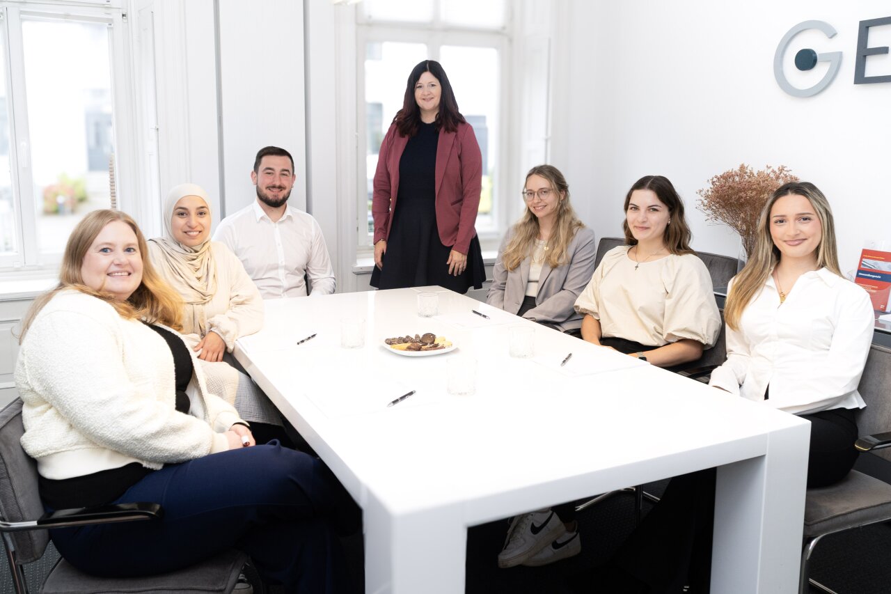 Eine Gruppe von sieben EGIDO-Mitarbeitern sitzt rund um einen weißen Konferenztisch in einem hellen, modernen Raum. Im Hintergrund steht eine Frau in einem langen schwarzen Kleid mit einer roten Jacke, während die anderen sitzend teilweise lächeln und Lookbilder präsentieren. Auf dem Tisch stehen Gläser und ein Teller mit Snacks.
