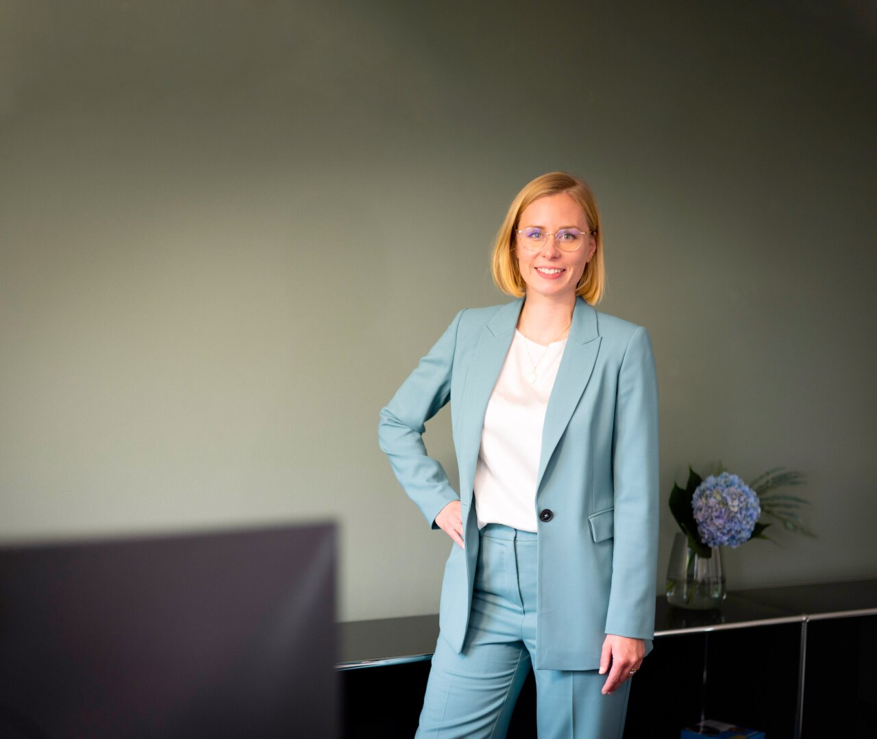 Eine lächelnde EGIDO-Mitarbeiterin mit blonden, schulterlangen Haaren trägt einen hellblauen Anzug und steht entspannt mit einer Hand in die Hüfte gestützt. Im Hintergrund sind eine graue Wand und eine Blumenvase mit blauen Blumen zu sehen.